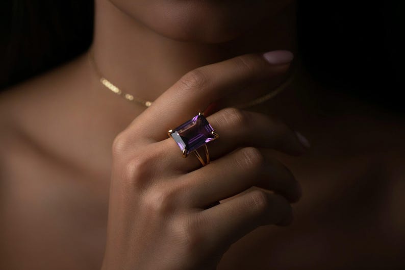 May include: A gold ring featuring a large, rectangular, purple amethyst gemstone. The ring is set on a gold band with a split shank design. The ring is worn on a hand, with a gold necklace visible in the background.