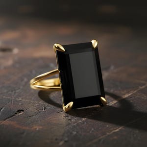 Puede incluir: Un anillo de oro con una gran piedra preciosa negra rectangular. El anillo tiene un dise&ntilde;o de ca&ntilde;a dividida y puntas doradas que aseguran la piedra. El engaste y la banda son de color oro pulido, creando un contraste llamativo.