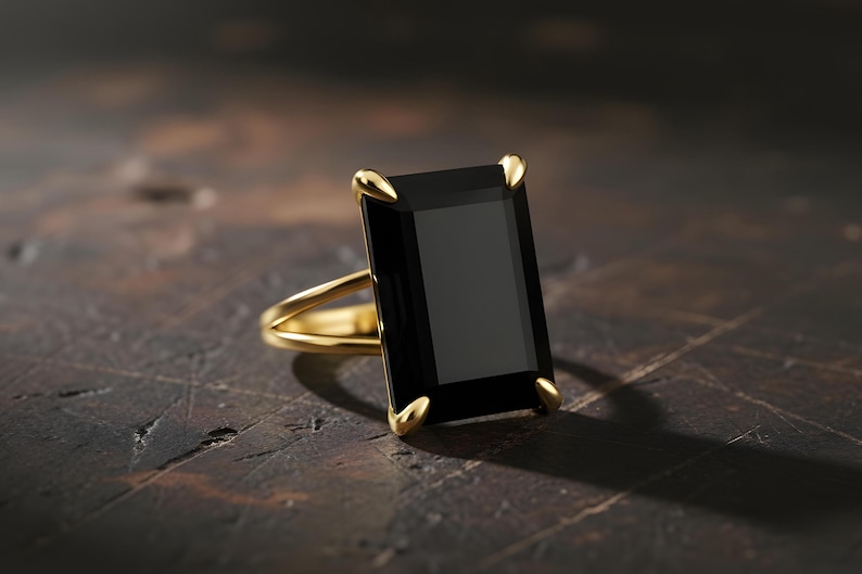 Puede incluir: Un anillo de oro con una gran piedra preciosa negra rectangular. El anillo tiene un dise&ntilde;o de ca&ntilde;a dividida y puntas doradas que aseguran la piedra. El engaste y la banda son de color oro pulido, creando un contraste llamativo.