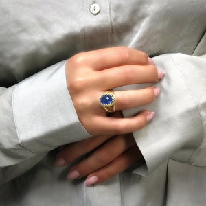 K&ouml;nnte beinhalten: Ein goldener Ring mit einem gro&szlig;en blauen ovalen Edelstein, der in eine strukturierte Goldband gefasst ist. Der Ring wird am Zeigefinger einer Hand getragen.