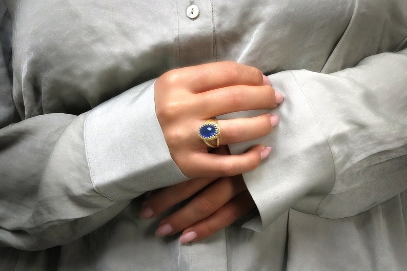 K&ouml;nnte beinhalten: Ein goldener Ring mit einem gro&szlig;en blauen ovalen Edelstein, der in eine strukturierte Goldband gefasst ist. Der Ring wird am Zeigefinger einer Hand getragen.