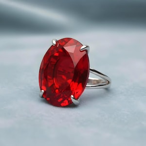 May include: A silver ring featuring a large, oval-cut, faceted red gemstone. The gemstone is held in place by four silver prongs. The ring is set against a soft, light blue background, highlighting the vibrant red color of the stone.