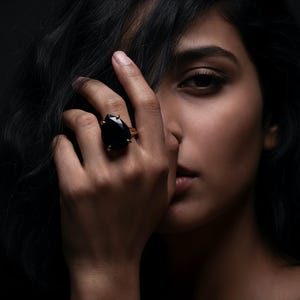 May include: A close-up of a hand wearing a large black gemstone ring with a gold setting. The ring is on the ring finger. The person's face is partially visible, with dark hair and a dark background.