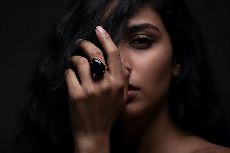 May include: A close-up of a hand wearing a large black gemstone ring with a gold setting. The ring is on the ring finger. The person's face is partially visible, with dark hair and a dark background.