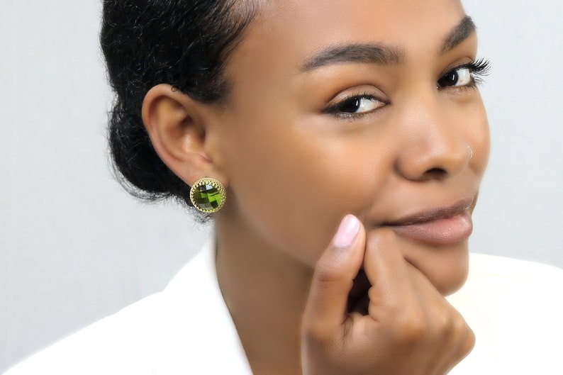 May include: A pair of gold earrings with a round green gemstone in the center. The earrings have a delicate, intricate design around the gemstone.