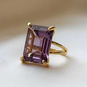May include: A gold ring featuring a large, rectangular, purple gemstone. The gemstone is set with four gold prongs. The ring's band splits into two strands as it approaches the gemstone. The ring is set against a white background.