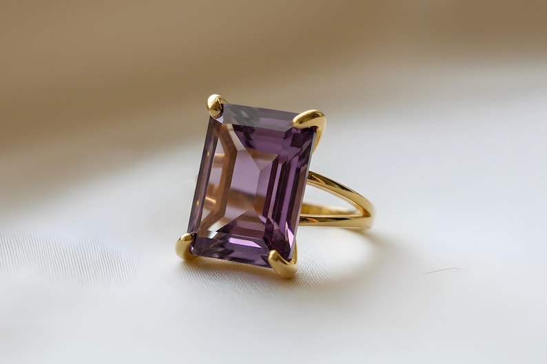 May include: A gold ring featuring a large, rectangular, purple gemstone. The gemstone is set with four gold prongs. The ring's band splits into two strands as it approaches the gemstone. The ring is set against a white background.