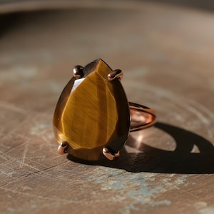 A large, irregularly shaped amber-colored gemstone, possibly a diamond or amber, is displayed on a metal ring against a textured background.