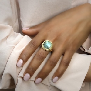 Puede incluir: Un anillo de oro con una gran piedra preciosa verde de forma ovalada engastada en un engaste de cuentas. El anillo está en un dedo.