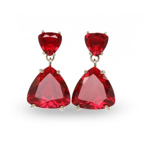 Puede incluir: Un par de pendientes colgantes con gemas rojas triangulares vibrantes. Cada pendiente tiene una piedra más pequeña en forma de corazón en la parte superior, conectada por una cadena plateada a una piedra triangular más grande debajo.