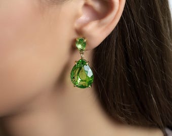 Pode incluir: Close-up de um brinco dourado com uma pedra preciosa verde brilhante em forma de pêra. O brinco tem uma pedra verde redonda menor na parte superior. A joia é colocada em um fundo desfocado de pele e cabelo.