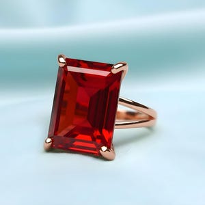 May include: A rose gold ring featuring a large, rectangular, deep red gemstone. The ring has a split shank design and is set against a soft, light blue background. The gemstone is faceted, catching the light.