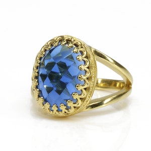 K&ouml;nnte beinhalten: Ein goldener Ring mit einem gro&szlig;en, facettierten blauen Edelstein, der in einer verzierten goldenen Fassung sitzt.