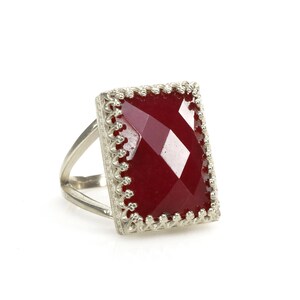 May include: A silver ring with a large, rectangular, faceted, deep red gemstone. The gemstone is set in a decorative, filigree-style bezel. The ring has a split shank design.