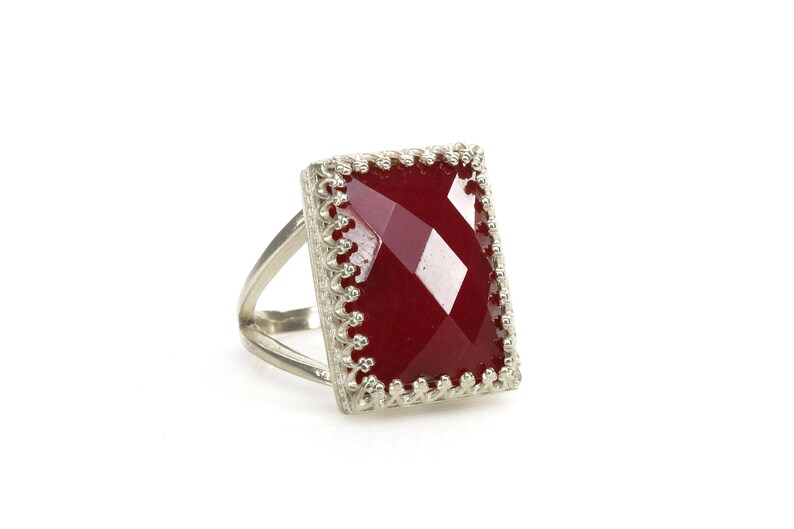 May include: A silver ring with a large, rectangular, faceted, deep red gemstone. The gemstone is set in a decorative, filigree-style bezel. The ring has a split shank design.