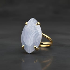 May include: A gold ring featuring a large, marquise-cut, pale blue gemstone with white veining. The ring has a split shank design and gold prongs securing the stone. The ring is set against a dark gray background.