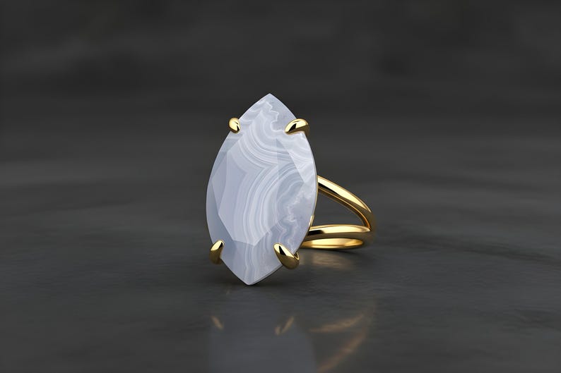 May include: A gold ring featuring a large, marquise-cut, pale blue gemstone with white veining. The ring has a split shank design and gold prongs securing the stone. The ring is set against a dark gray background.