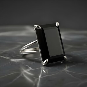 Puede incluir: Un anillo de plata con una gran gema negra rectangular. El anillo tiene un dise&ntilde;o de ca&ntilde;a dividida y est&aacute; colocado sobre una superficie oscura y veteada. La gema est&aacute; sujeta por cuatro garras de plata.