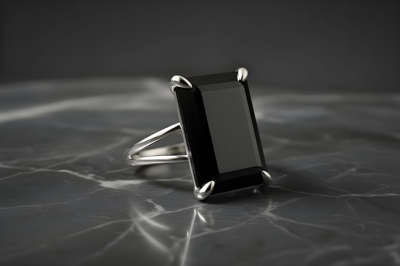 Puede incluir: Un anillo de plata con una gran gema negra rectangular. El anillo tiene un dise&ntilde;o de ca&ntilde;a dividida y est&aacute; colocado sobre una superficie oscura y veteada. La gema est&aacute; sujeta por cuatro garras de plata.
