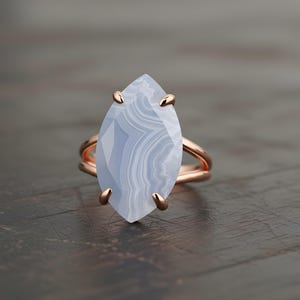 May include: A rose gold ring featuring a large, marquise-cut blue lace agate gemstone. The stone has a layered, banded appearance. The ring is set on a dark wooden surface.