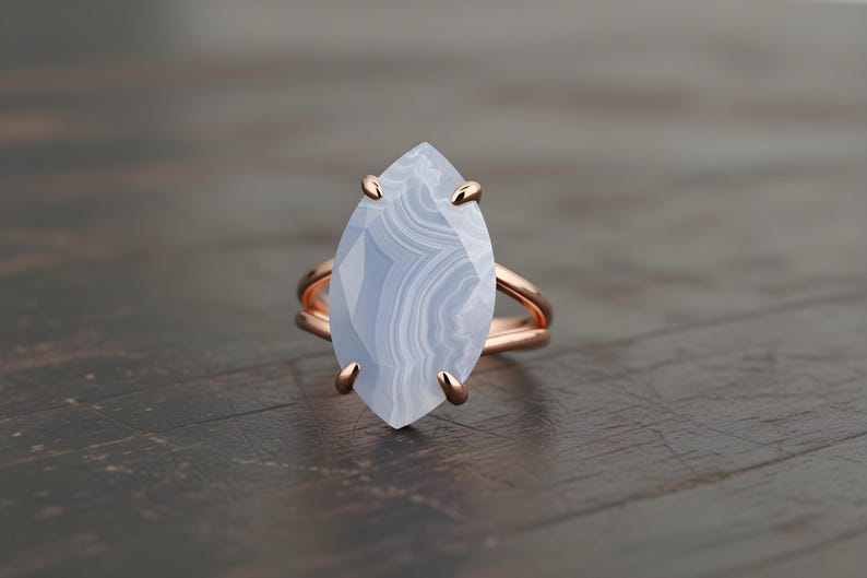 May include: A rose gold ring featuring a large, marquise-cut blue lace agate gemstone. The stone has a layered, banded appearance. The ring is set on a dark wooden surface.