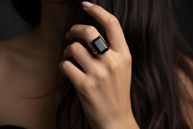 Puede incluir: Un anillo dorado con una gran piedra negra rectangular. El anillo tiene un dise&ntilde;o minimalista, con la piedra negra como elemento principal. La gema refleja la luz, creando un efecto llamativo. El anillo se lleva en una mano, con cabello oscuro en el fondo.