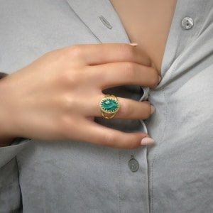 May include: Gold ring featuring an oval, green gemstone with a textured surface. The ring has a detailed, openwork design around the gemstone and a delicate band. The ring is worn on a finger.