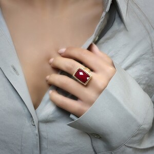 May include: A gold-toned ring with a large, rectangular, deep red gemstone. The ring has a textured, ornate border. The ring is worn on a finger, with a gray button-down shirt in the background.