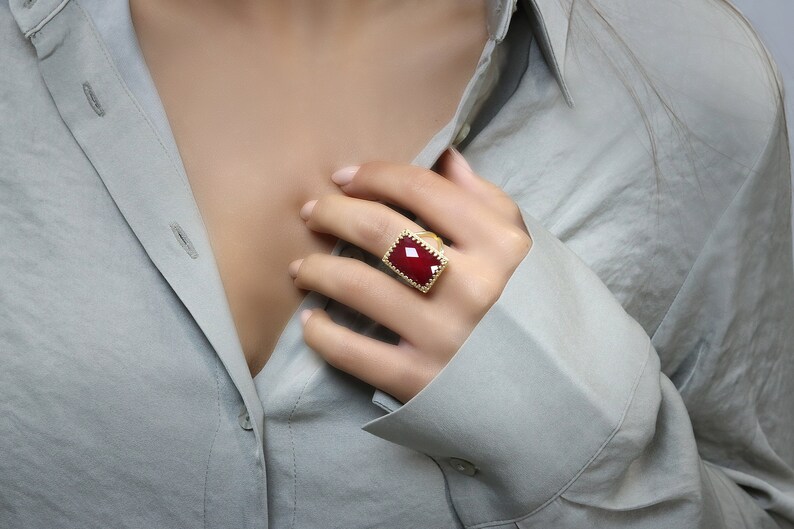 May include: A gold-toned ring with a large, rectangular, deep red gemstone. The ring has a textured, ornate border. The ring is worn on a finger, with a gray button-down shirt in the background.