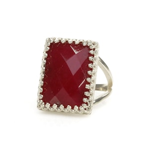 May include: A silver ring with a large, rectangular, faceted, deep red gemstone. The gemstone is set in a decorative, scalloped bezel. The ring band is a simple, split design.