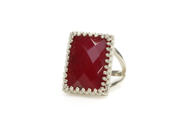 May include: A silver ring with a large, rectangular, faceted, deep red gemstone. The gemstone is set in a decorative, scalloped bezel. The ring band is a simple, split design.