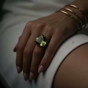 Könnte beinhalten: Ein goldener Ring mit einem großen, dreieckigen, grünen Edelstein. Der Ring befindet sich an einer Hand mit mehreren goldenen Armreifen am Handgelenk. Der Hintergrund ist eine weiche, neutrale Farbe.