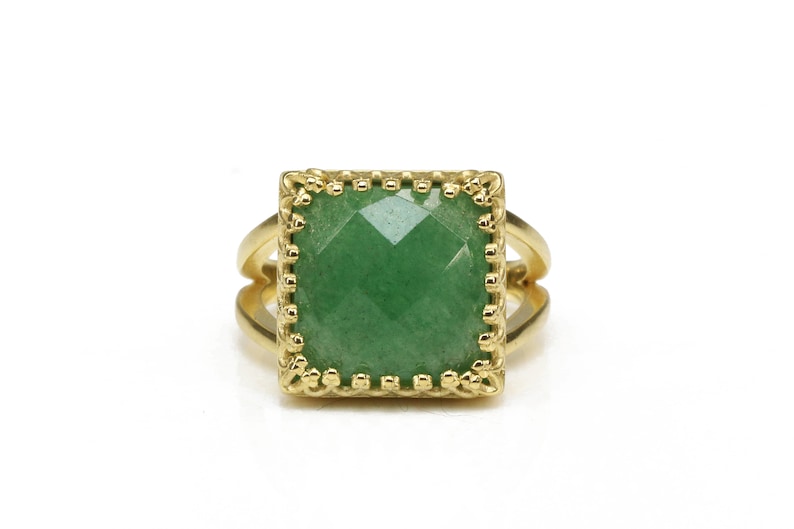 Puede incluir: Anillo dorado con una piedra preciosa verde cuadrada y facetada. La piedra preciosa est&aacute; engastada en un bisel decorativo y festoneado. El anillo tiene un dise&ntilde;o de ca&ntilde;a dividida, con dos bandas a cada lado de la piedra.