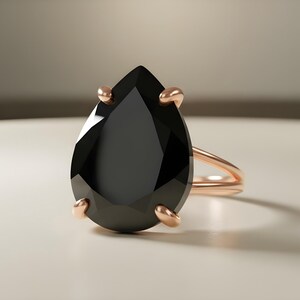 May include: A rose gold ring featuring a large, pear-shaped black gemstone. The gemstone is held in place by four prongs. The ring band is a simple, thin design. The ring is set against a neutral background.