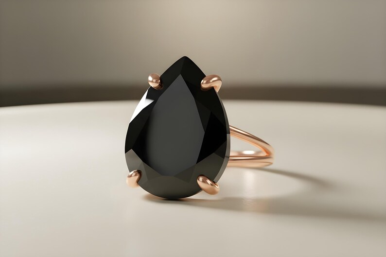 May include: A rose gold ring featuring a large, pear-shaped black gemstone. The gemstone is held in place by four prongs. The ring band is a simple, thin design. The ring is set against a neutral background.