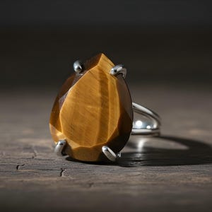 A large, irregularly shaped amber-colored gemstone, possibly a tiger's eye, is displayed on a silver ring against a dark background.
