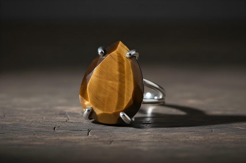 A large, irregularly shaped amber-colored gemstone, possibly a tiger's eye, is displayed on a silver ring against a dark background.