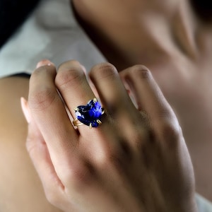 May include: A gold ring featuring a large, heart-shaped, deep blue gemstone. The gemstone is held in place by four gold prongs. The ring is on a person's finger, with the hand in the foreground.