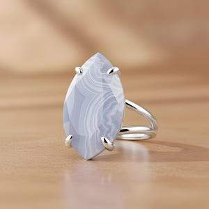 May include: A silver ring featuring a large, light blue gemstone with white marbling. The gemstone is marquise-shaped and held in place by four silver prongs. The ring has a split shank design.