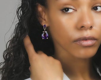 May include: A close-up of a person wearing a pair of drop earrings. The earrings feature a silver setting with two purple gemstones. The top stone is small and round, while the bottom stone is a larger, teardrop shape.