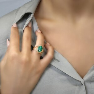 May include: A gold ring featuring a large, oval-shaped green stone. The ring has a detailed, textured band. The ring is worn on a finger, with a light gray shirt in the background.