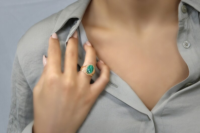 May include: A gold ring featuring a large, oval-shaped green stone. The ring has a detailed, textured band. The ring is worn on a finger, with a light gray shirt in the background.