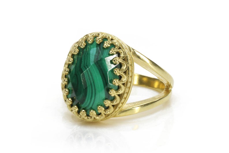 May include: A gold ring featuring a large, oval-cut malachite stone with a vibrant green color and intricate veining. The ring has a split shank design and a decorative, openwork bezel setting.