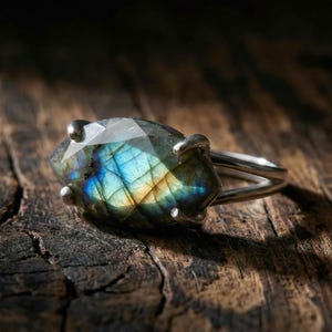 May include: A silver ring featuring a large, faceted labradorite gemstone. The stone displays iridescent blue, green, and yellow hues. The ring is set on a split shank band, resting on a textured wooden surface.