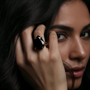 May include: A gold-toned ring featuring a large, black, teardrop-shaped gemstone. The ring is set with small gold prongs. The image is a close-up of the ring on a hand, with a dark background.