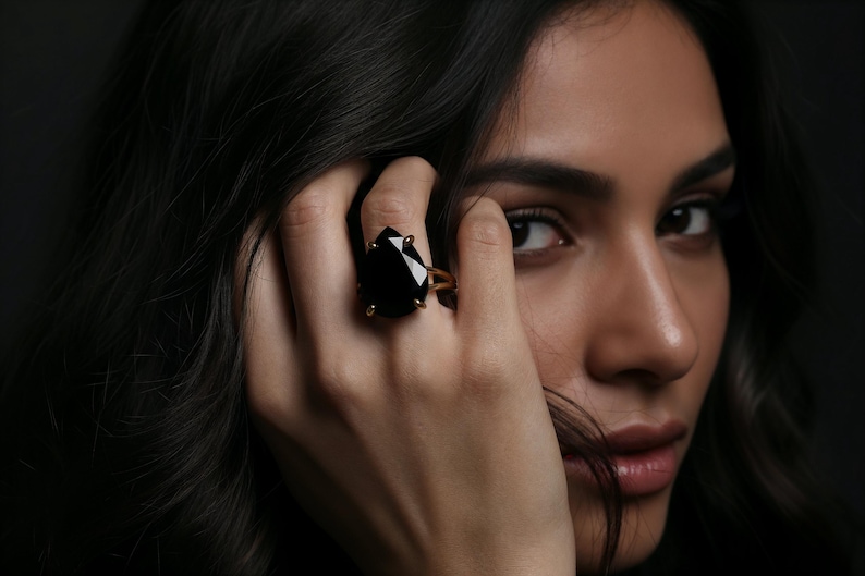 May include: A gold-toned ring featuring a large, black, teardrop-shaped gemstone. The ring is set with small gold prongs. The image is a close-up of the ring on a hand, with a dark background.