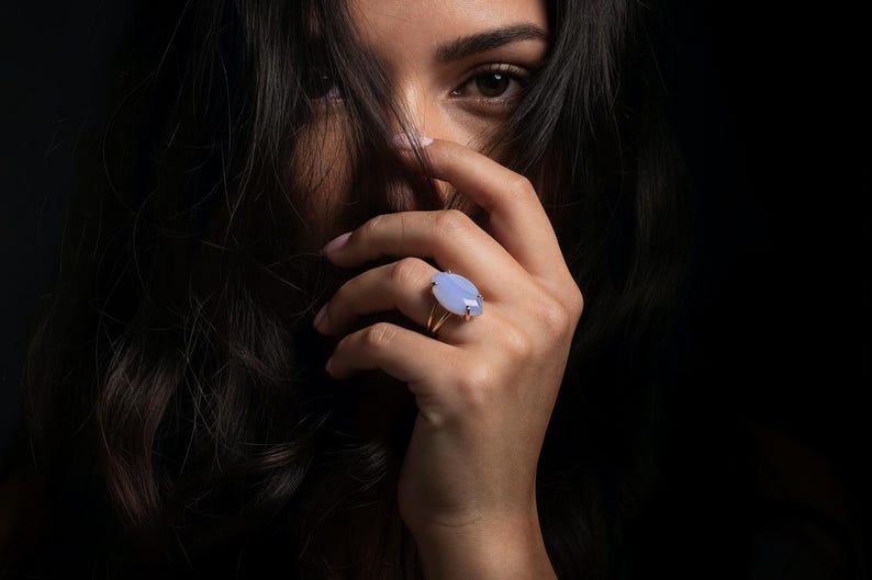 May include: A gold ring with a large, oval, pale blue gemstone. The ring is on a hand with long, dark hair partially obscuring the face. The background is dark.