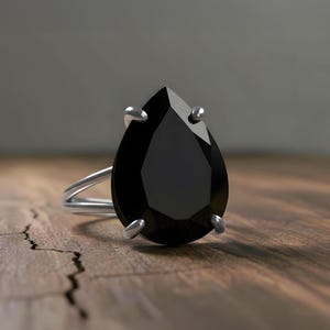 May include: A silver ring with a large, pear-shaped black gemstone. The ring's band is a double-loop design. The gemstone is held in place by four silver prongs. The ring is set on a wooden surface.