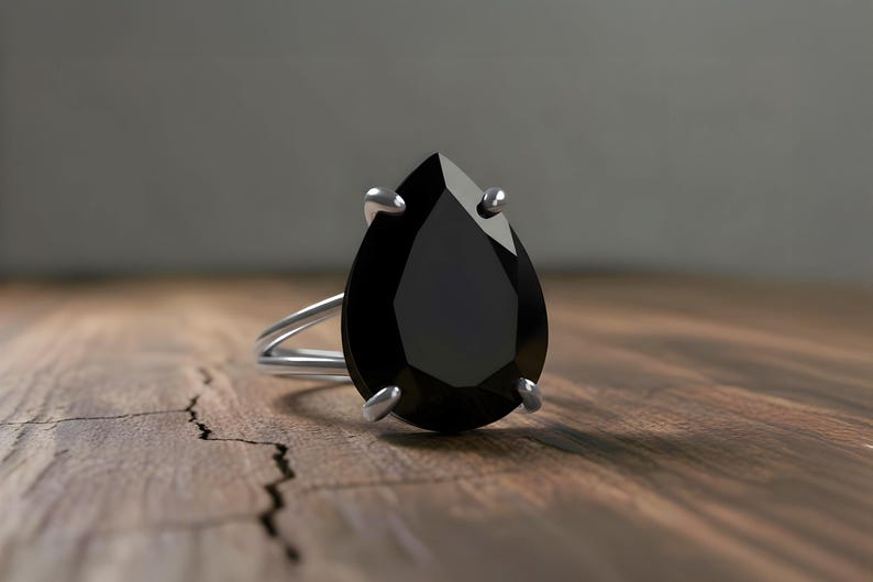 May include: A silver ring with a large, pear-shaped black gemstone. The ring's band is a double-loop design. The gemstone is held in place by four silver prongs. The ring is set on a wooden surface.