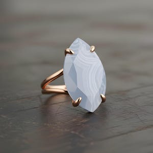 May include: A rose gold ring featuring a large, light blue gemstone with a unique, elongated shape. The gemstone has a marbled appearance. The ring is set on a dark wooden surface.